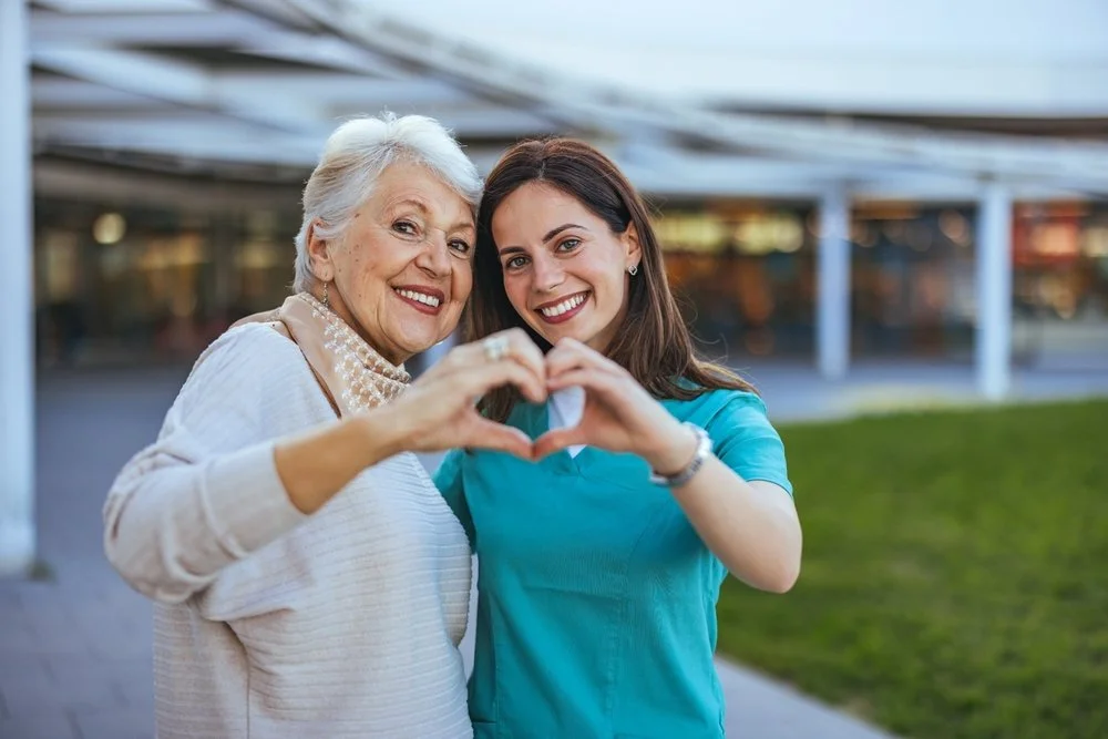 An older woman and a caregiver smiling and forming a heart with their hands