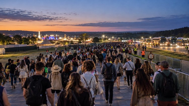 Illustrative festival crowd departing at dusk along a temporary route, with shuttle buses waiting alongside.