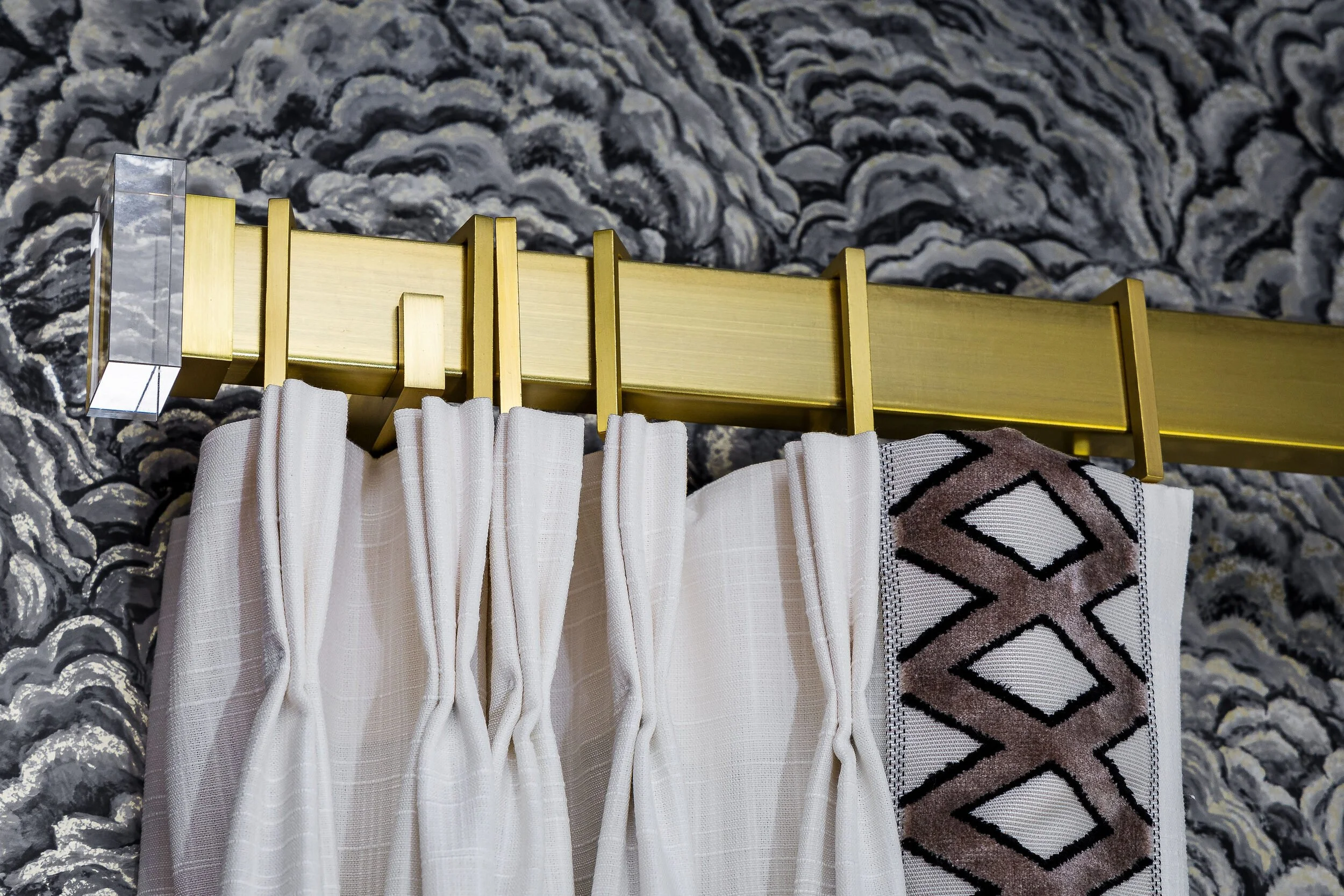 Detail of pleated drapery on a brass rod with a geometric trim against textured wallcovering