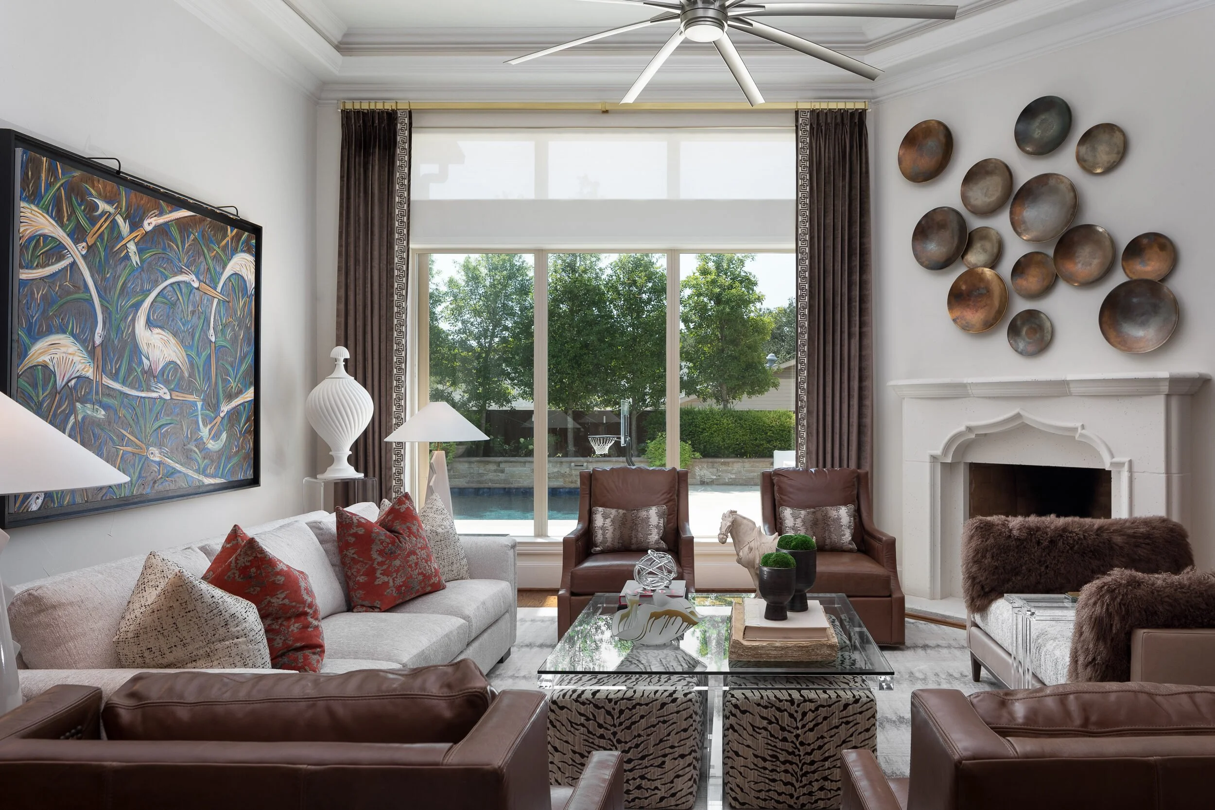 Living room with leather lounge chairs, a glass coffee table on patterned ottomans and a wall of bronze plates