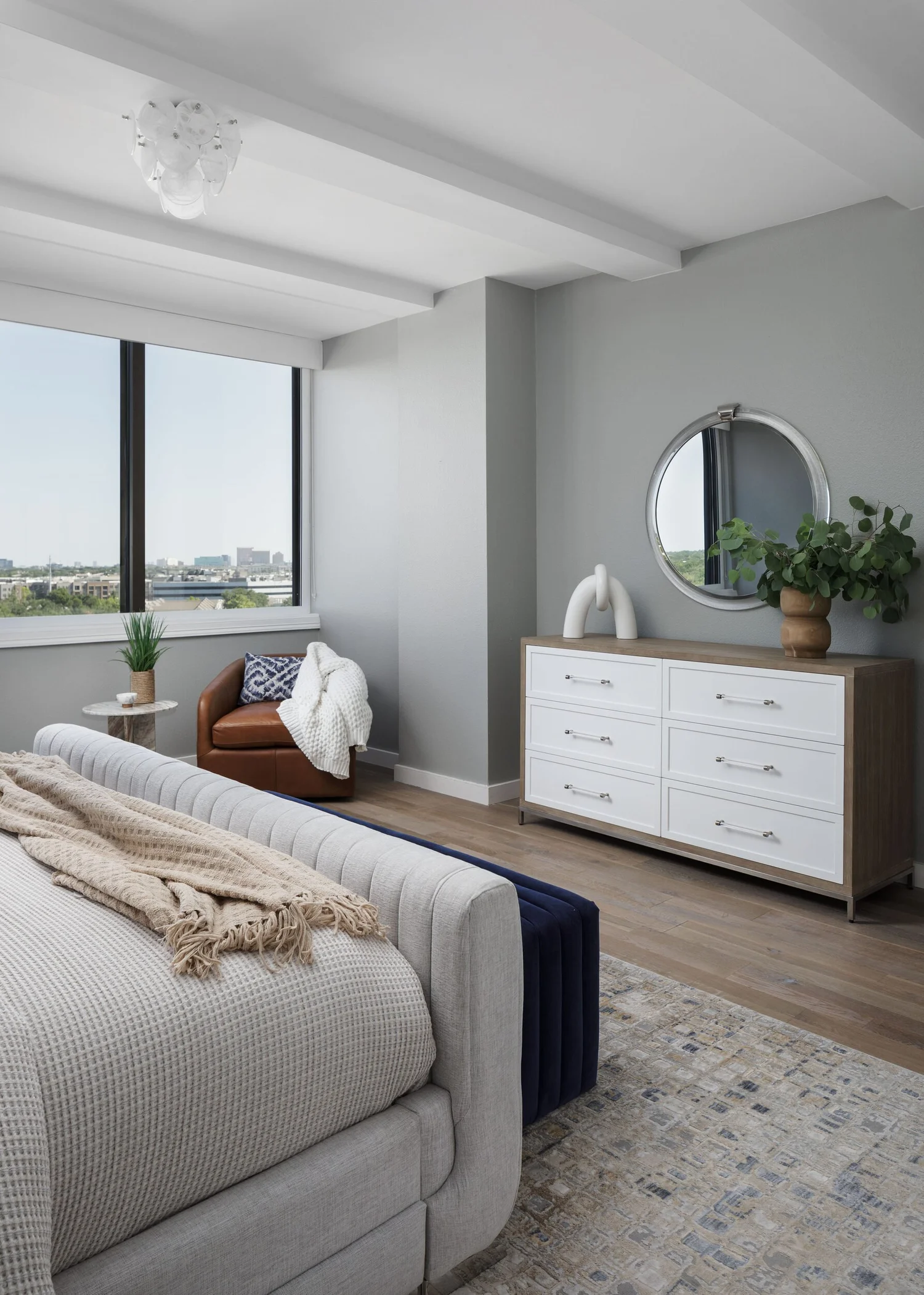 Bedroom corner with a white dresser, round mirror and city window