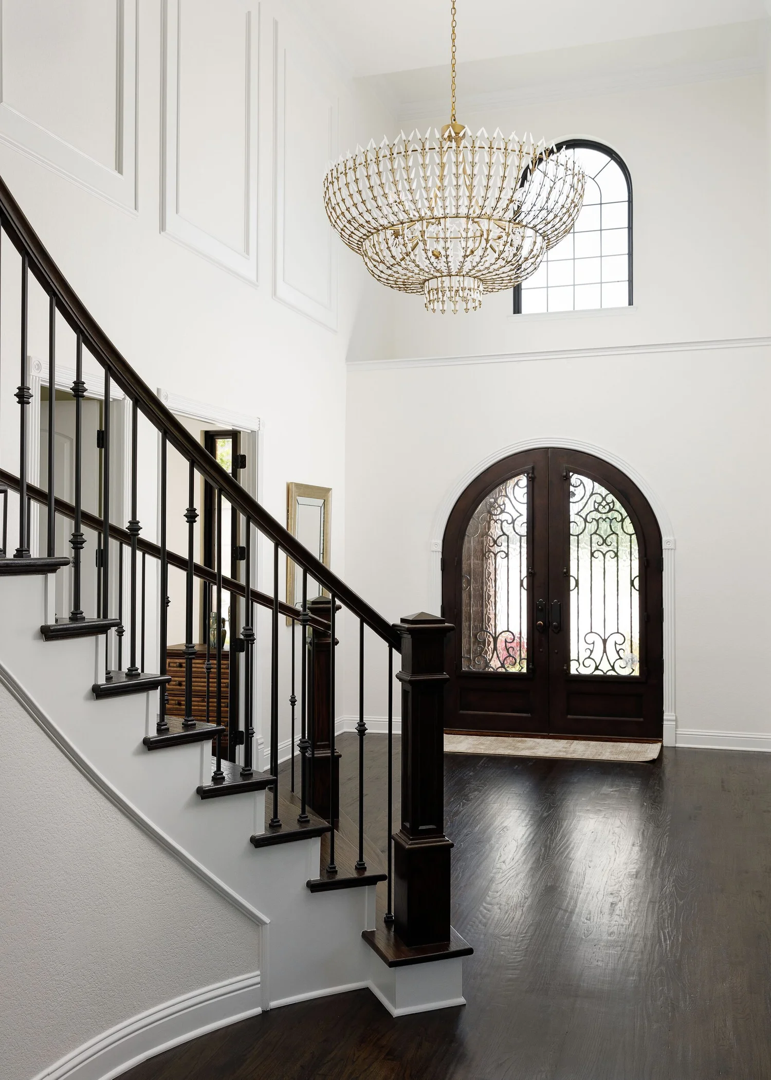 Curved staircase with an iron railing below a beaded chandelier and arched front doors