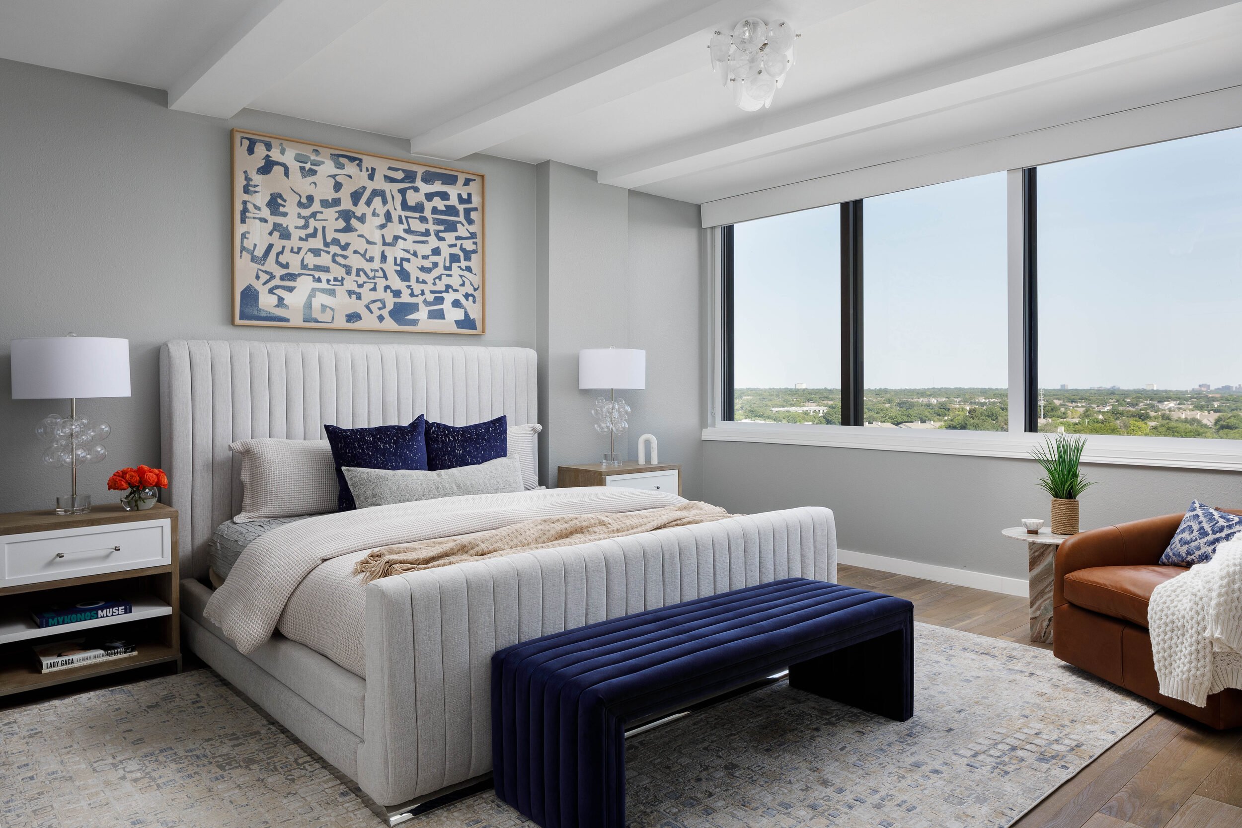 Primary bedroom with a channel-tufted bed, navy bench and a wall of windows over the city