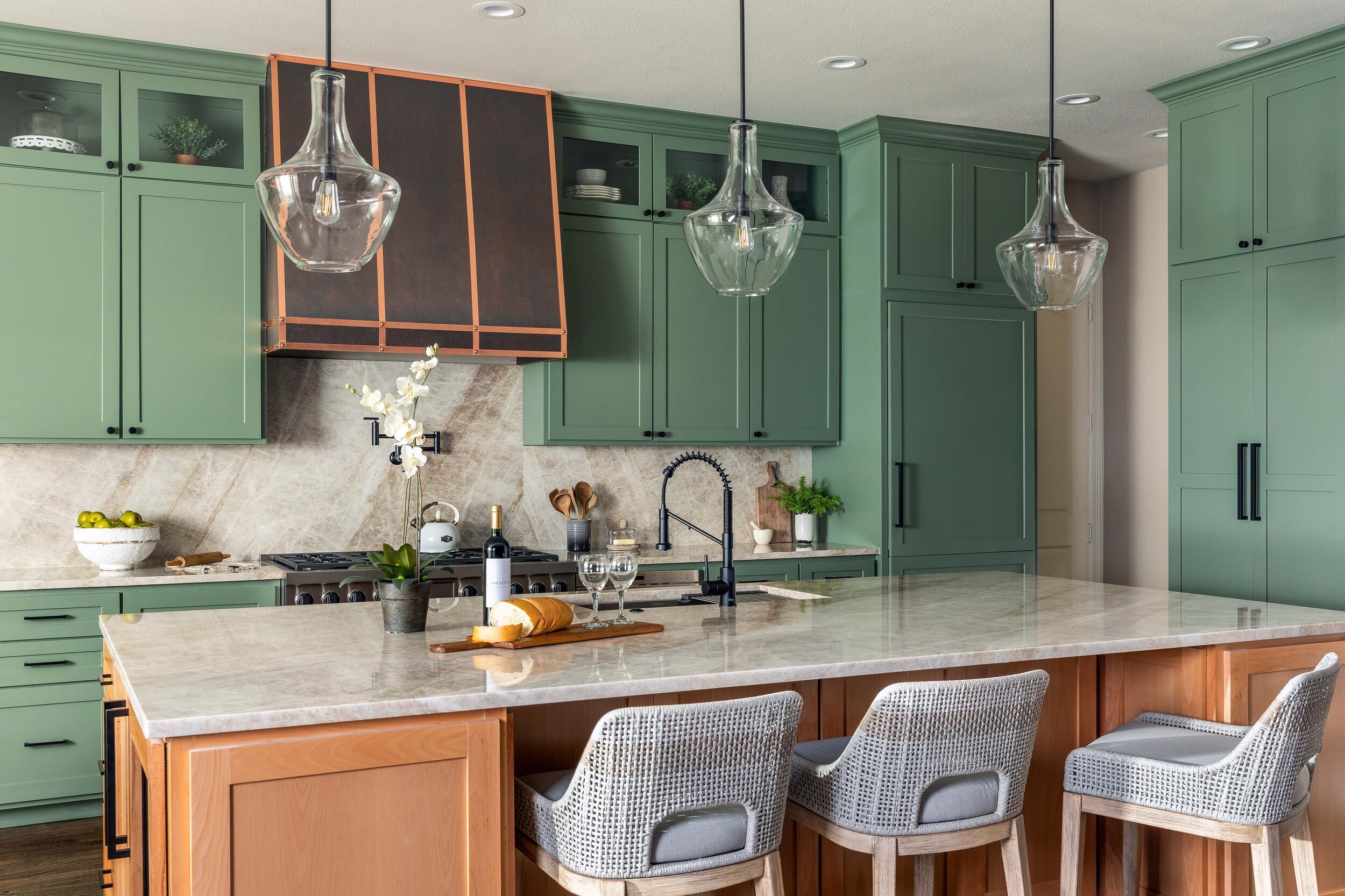 Island with woven counter stools beneath glass pendants, with the copper range hood beyond