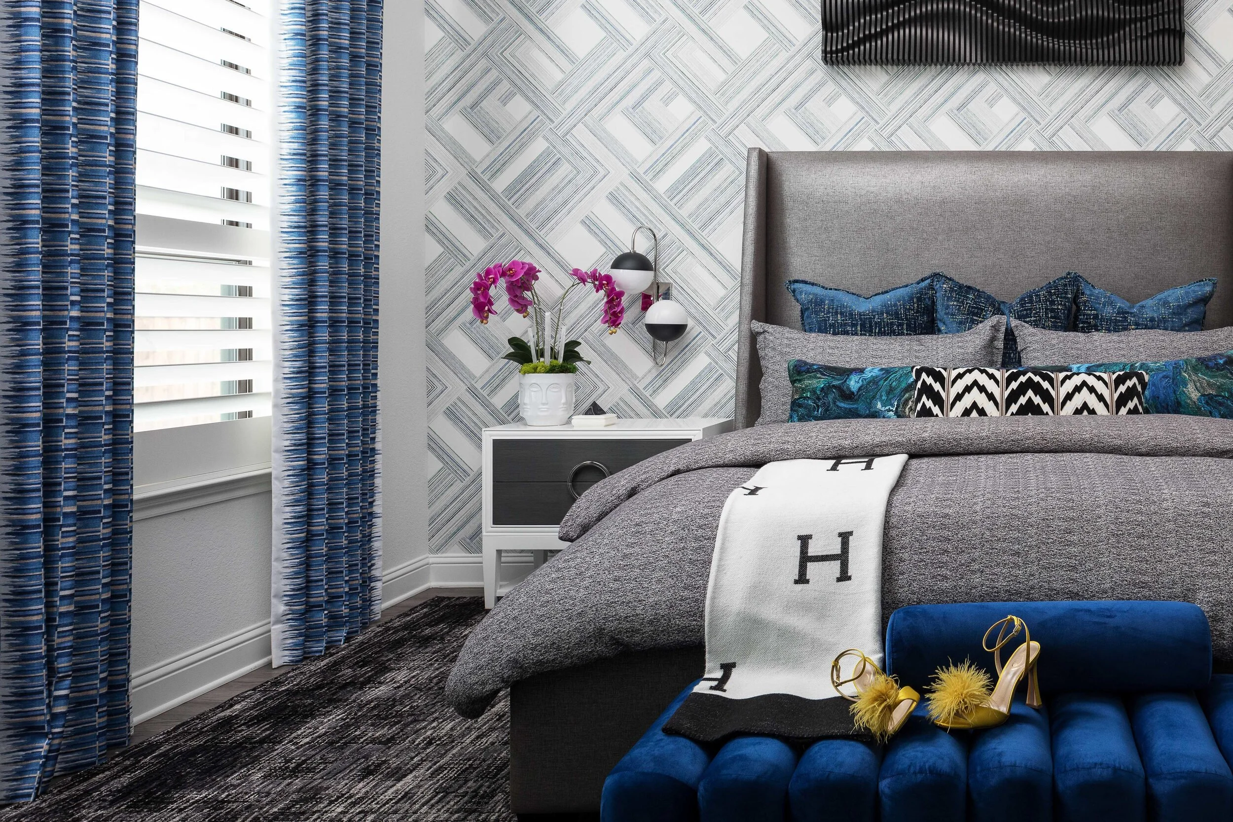 Primary bed with a blue tufted bench, orchids and a wall sconce against geometric wallpaper