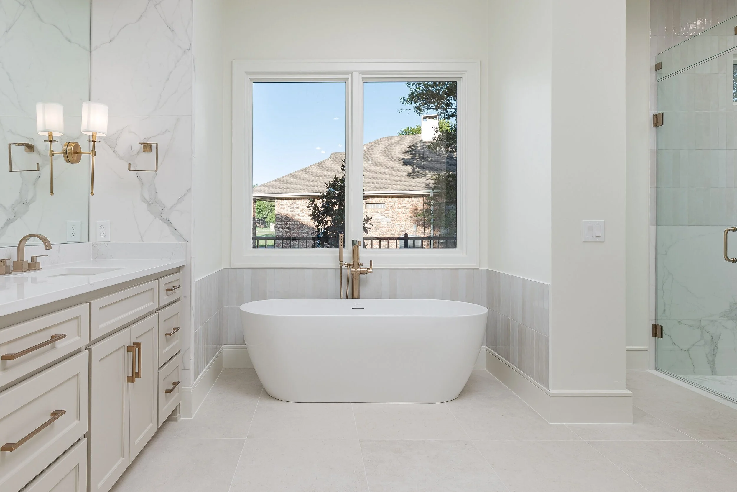 Freestanding tub centered beneath a double window between the vanity and the glass shower
