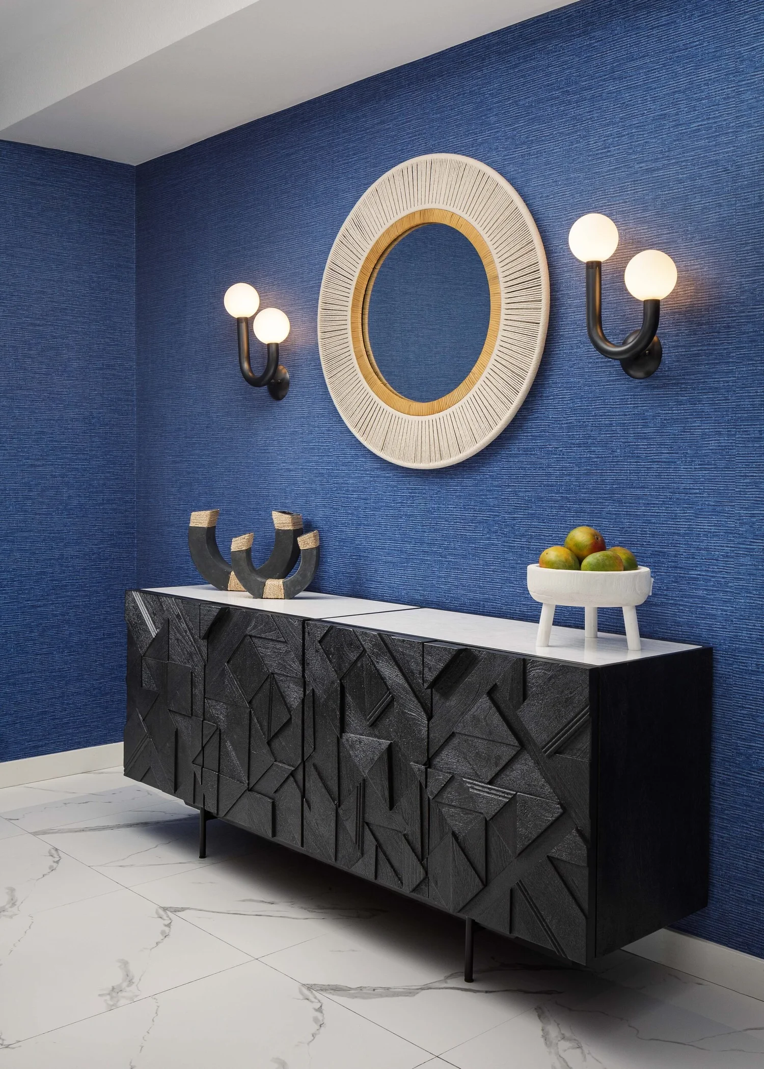 Black carved sideboard below a round rattan mirror and brass sconces on blue grasscloth