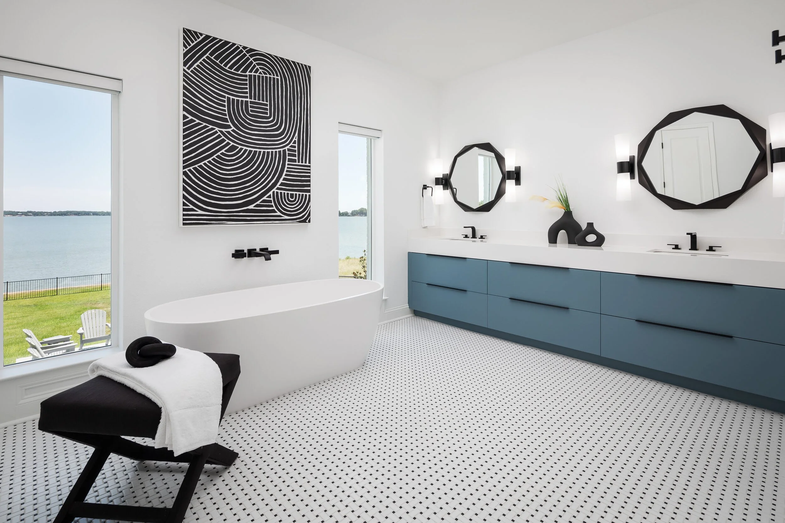 Freestanding tub beside lake-view windows, with the blue-gray vanity and black line art beyond