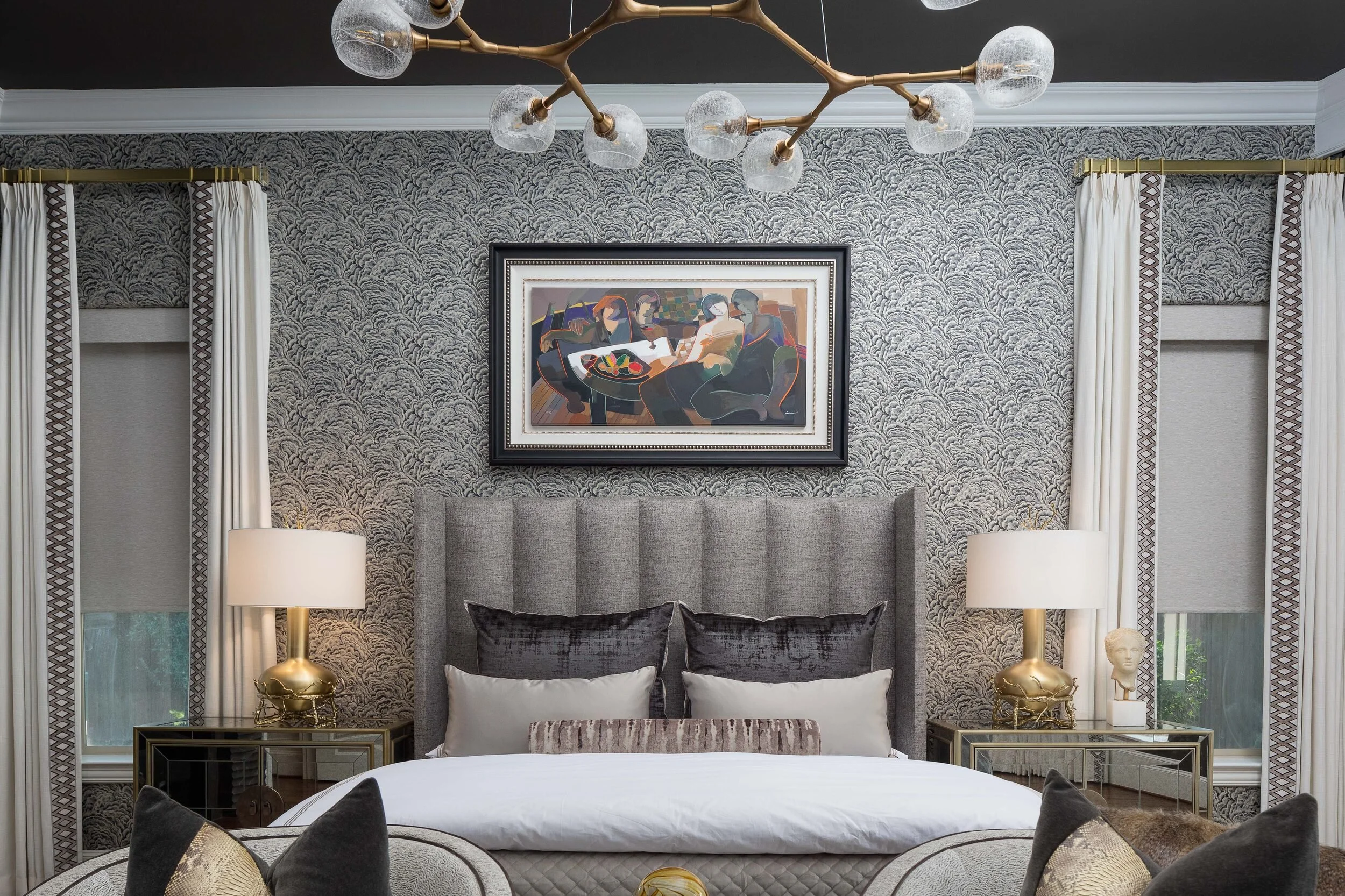 Straight-on view of a channel-tufted bed beneath framed art, with brass lamps and patterned wallcovering