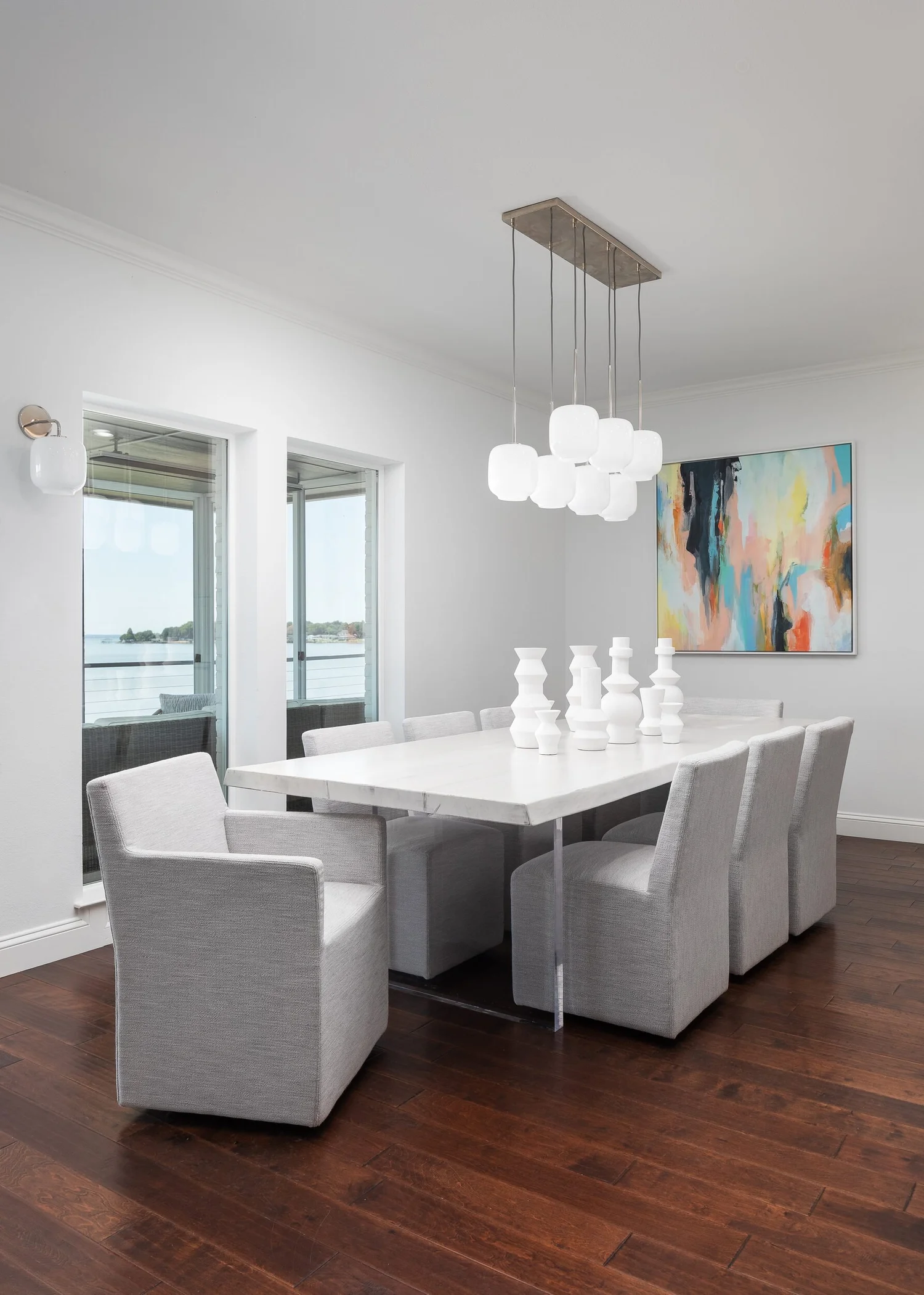 Dining room with a white table, upholstered chairs and a cluster pendant beside lake-view doors