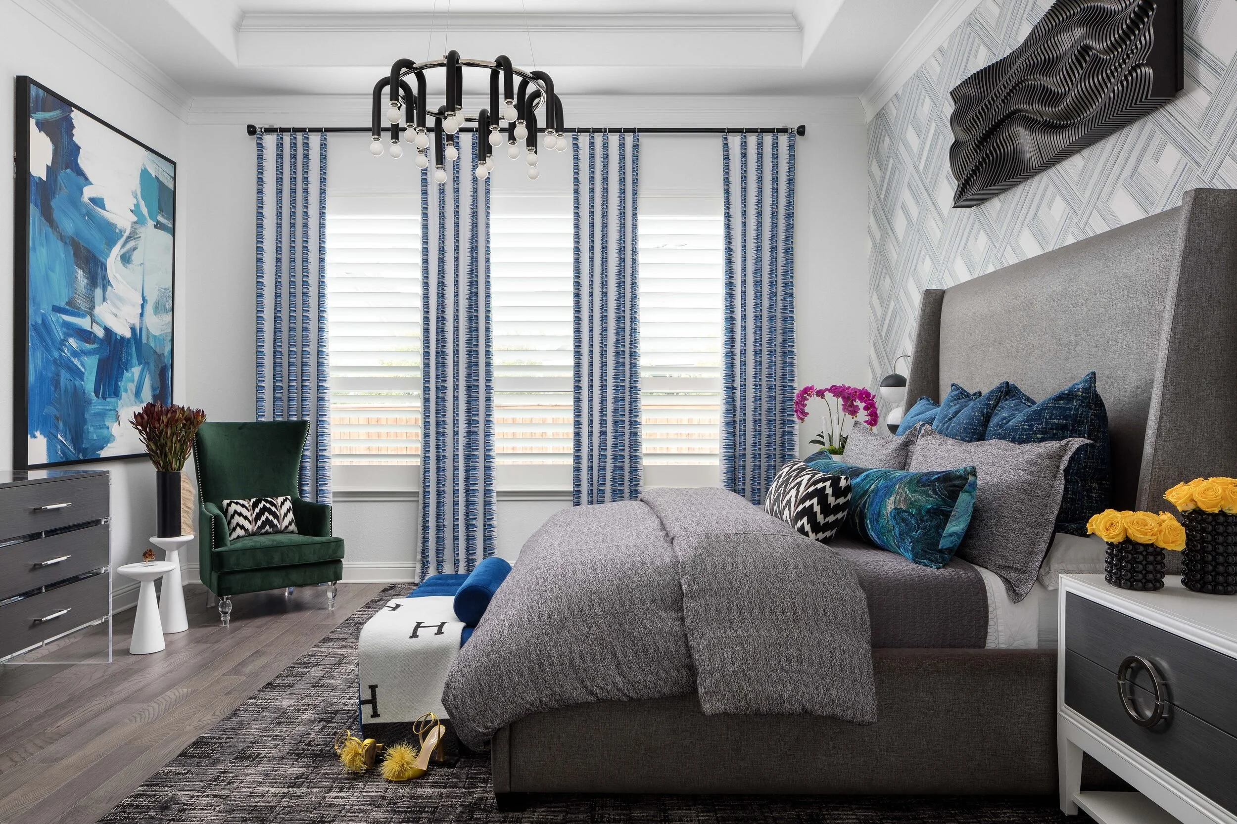 Primary bedroom with a gray wingback bed, geometric wallpaper, blue drapery and a black chandelier