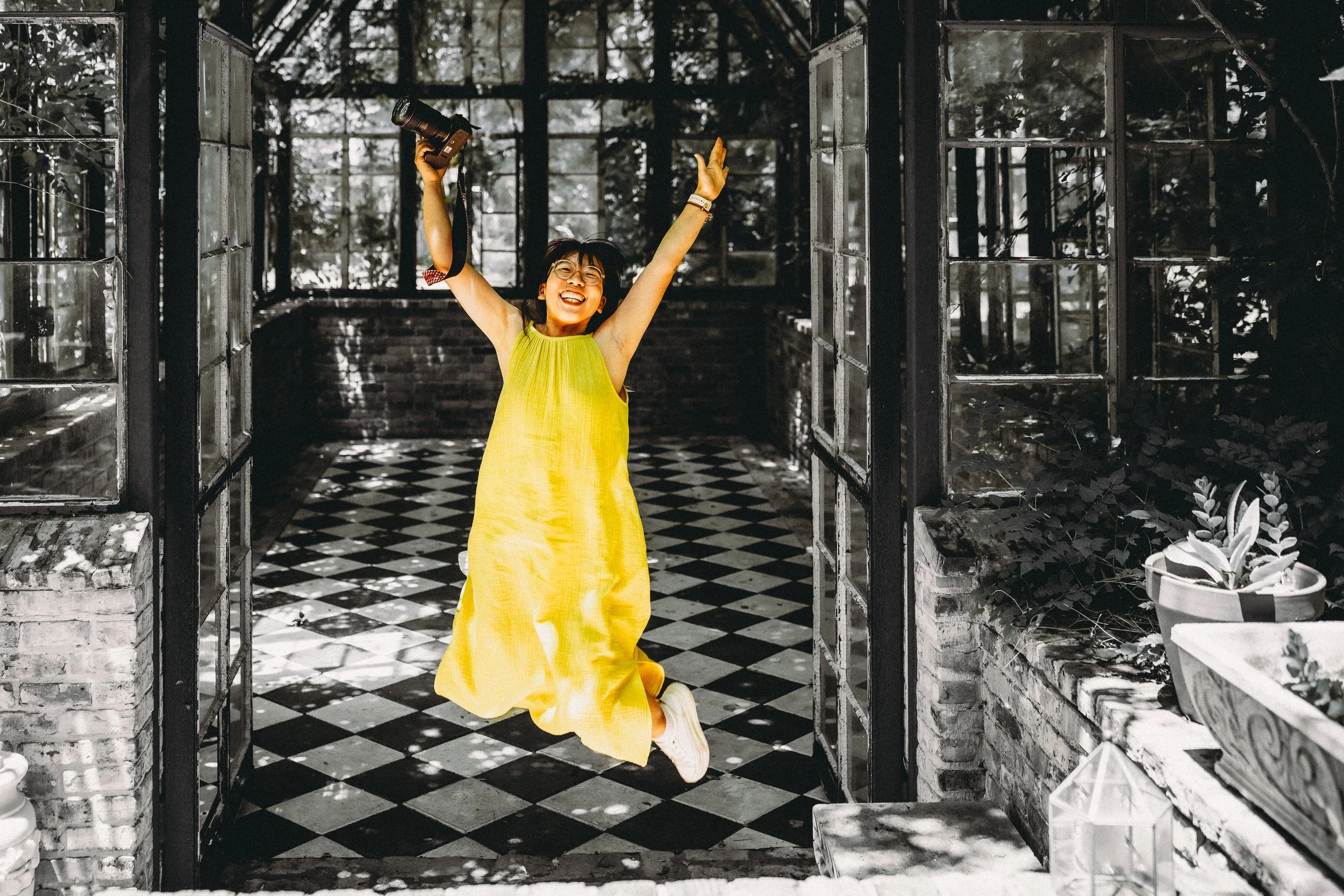 Aira Hickman, Maryland documentary wedding photographer, jumping joyfully with camera in a vintage greenhouse