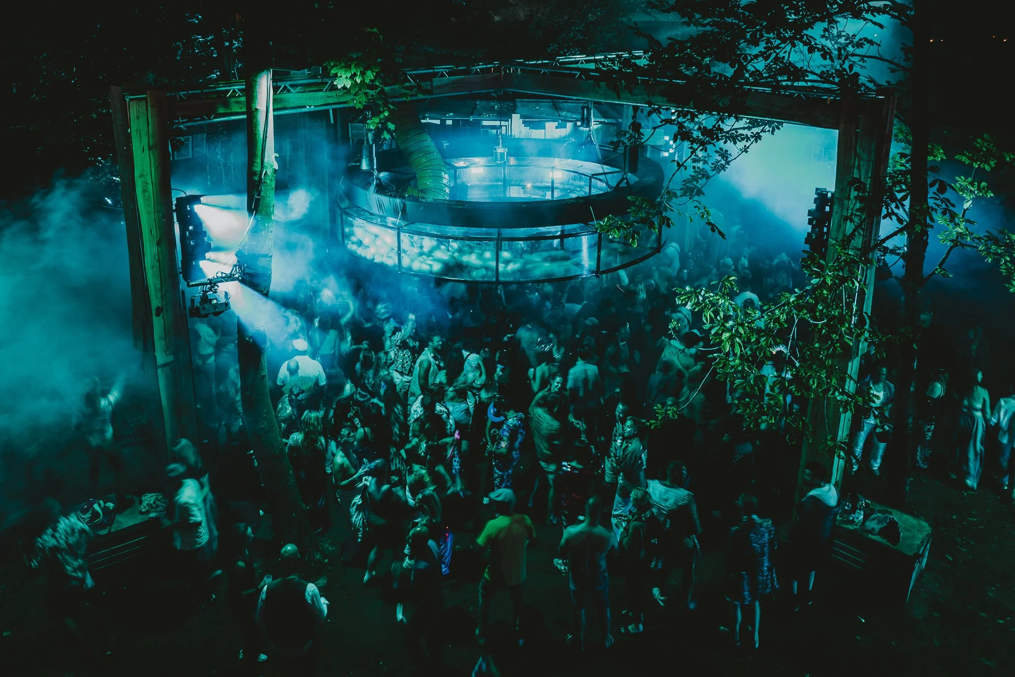 Stage design with a suspended circular installation, blue-green lighting and a crowd among the trees