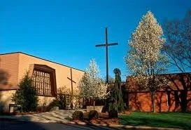 The Bergamo Center: a low brick building with a tall cross before it and white flowering trees, under a clear blue sky.