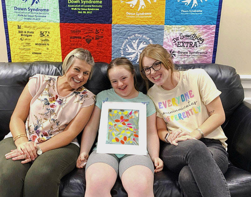 Three people smiling together on a couch while the person in the center holds framed artwork, with a DSACK quilt behind them
