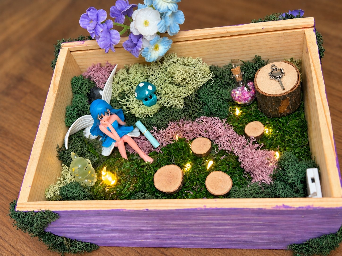 Finished fairy garden in a purple wooden box with moss, flowers, tiny mushrooms, and a fairy.