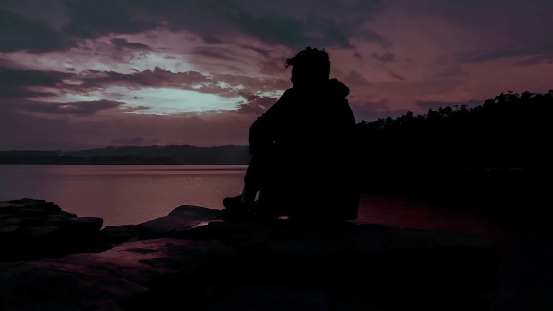A person sitting beside the water beneath a purple sunset sky
