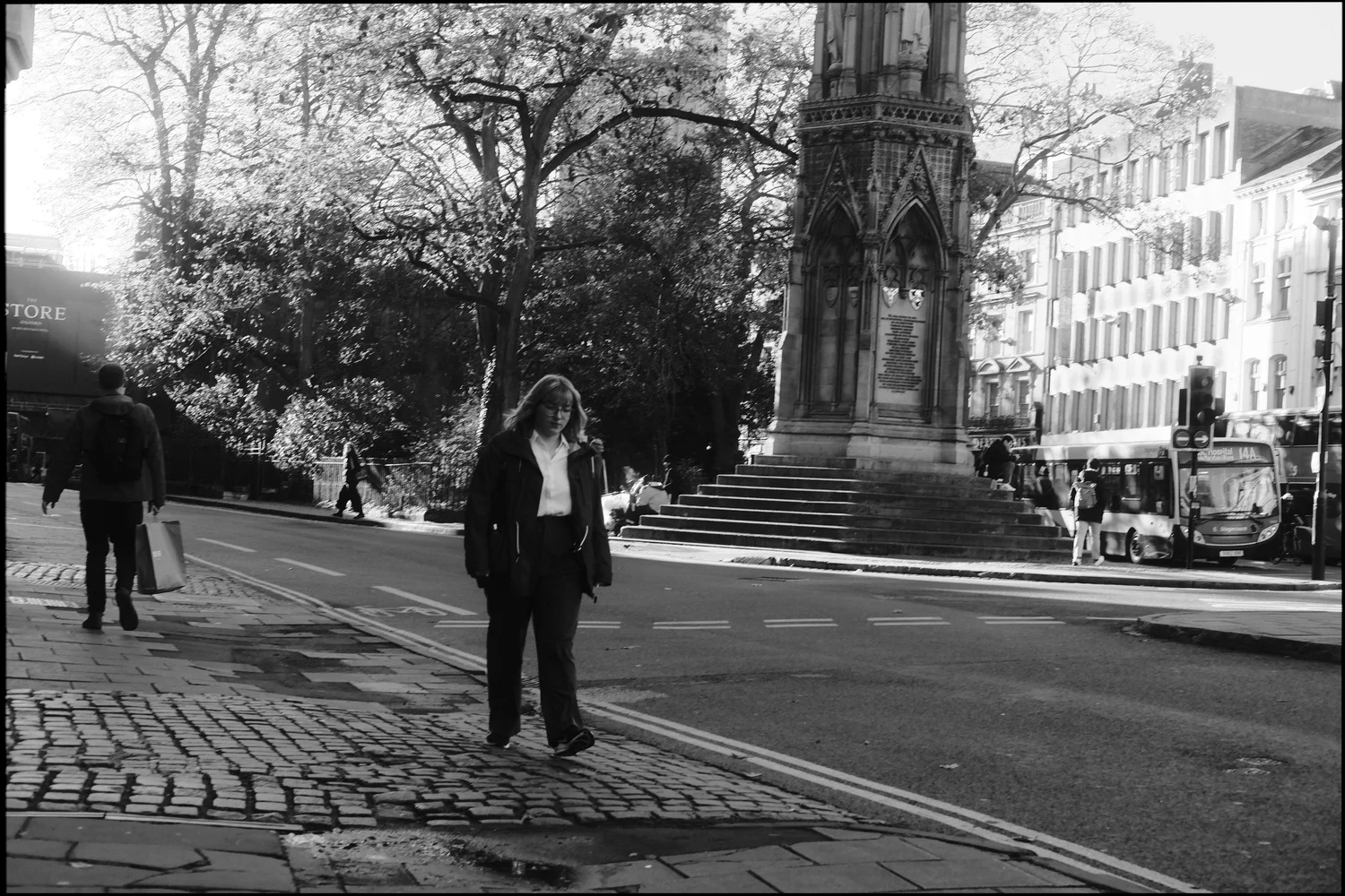 Black and white photo of a woman crossing a street toward the camera, with the Martyrs' Memorial and a bus behind her.