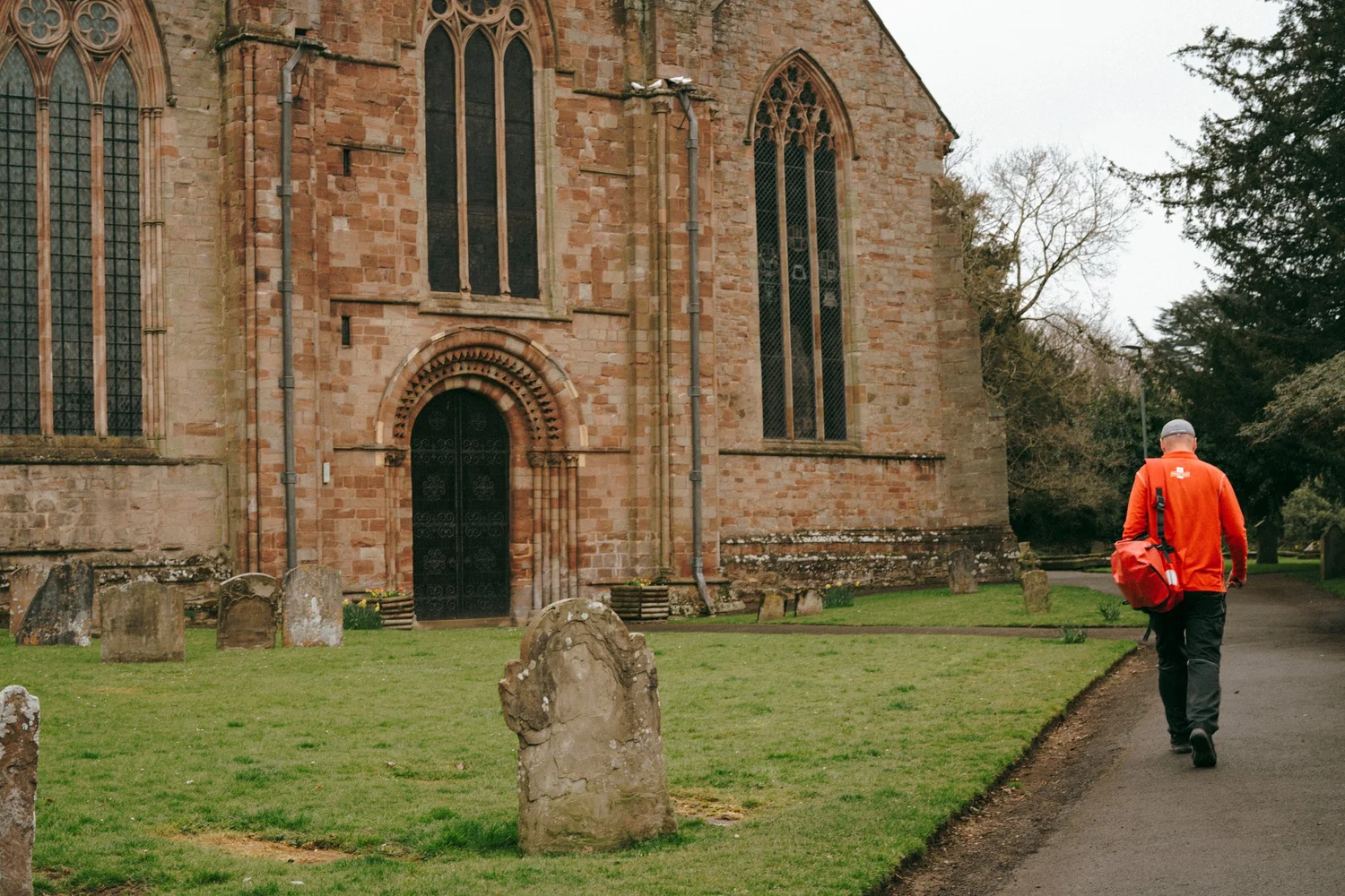 A hi-vis worker walking past a Norman-arched church door and gravestones.