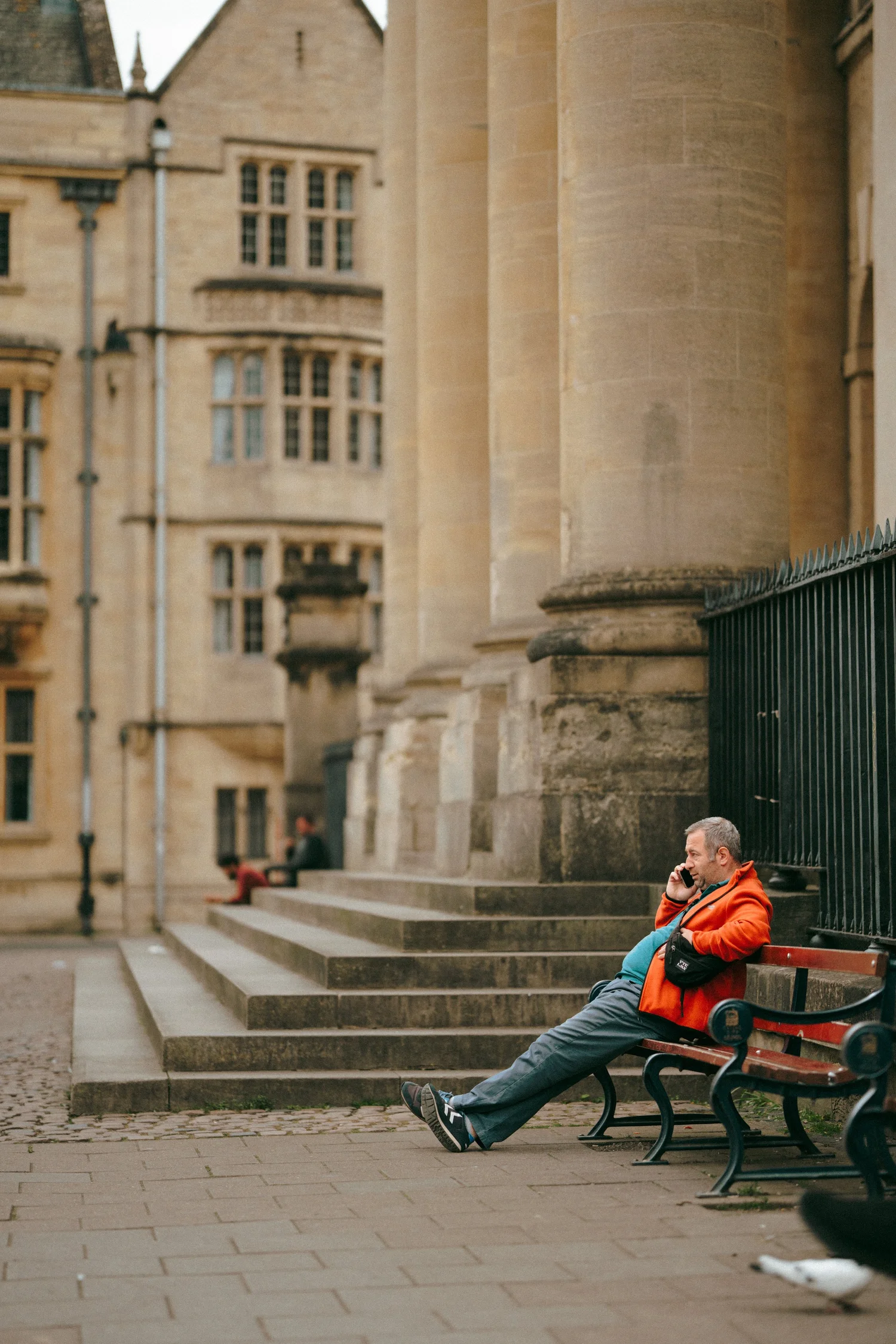A man in an orange coat reclining on a bench talking on the phone, in front of a grand college building.