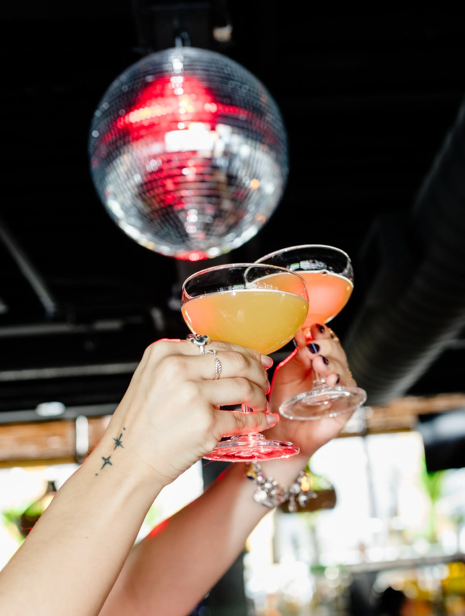 Two cocktails cheering in the air with a disco ball in the background inside at Avenida