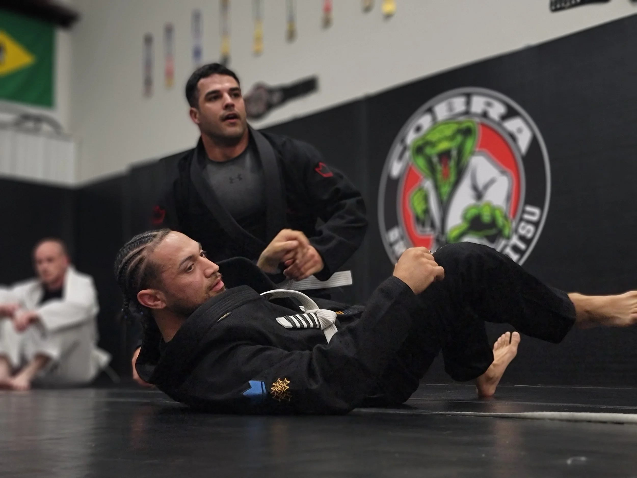 Two students practising a technique in front of the Cobra BJJ wall logo