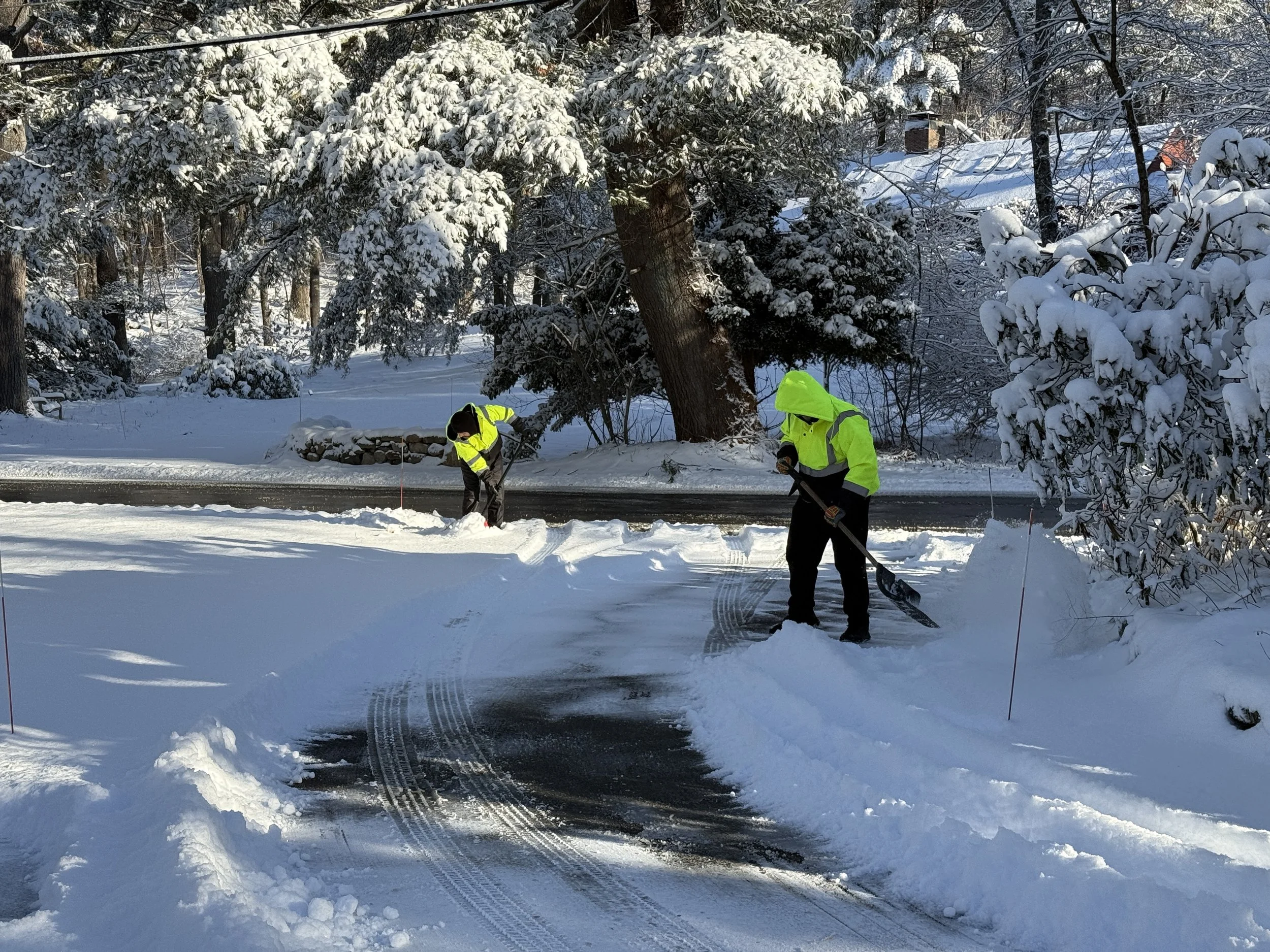 Two snow removal crew members in high-visibility jackets shoveling a residential driveway.
