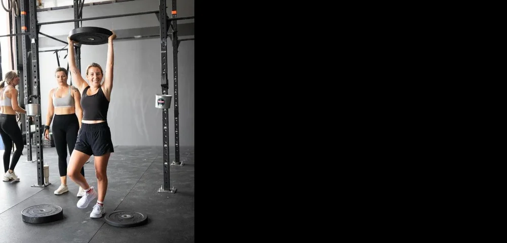 Member smiling while holding a bumper plate overhead during class