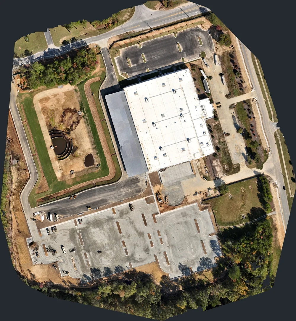 Aerial site mosaic combining overlapping construction photographs into an overhead view of the building, parking areas, access roads, and site basin.