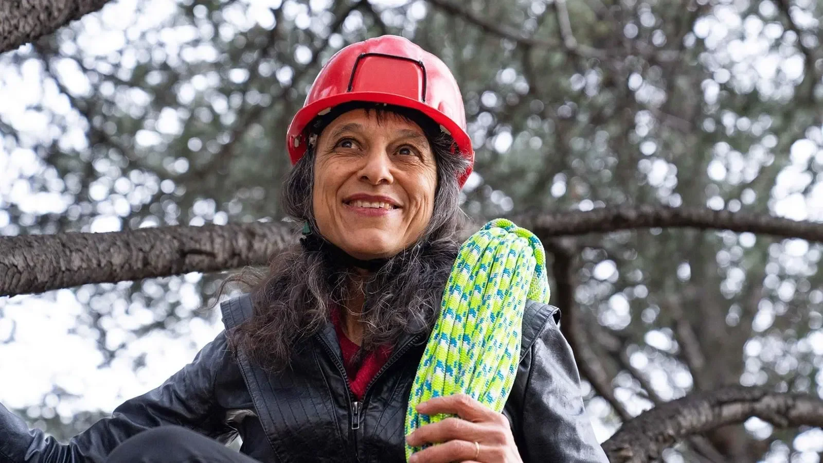 Dr. Nalini Nadkarni wears a red climbing helmet and holds a coiled rope among tree branches.