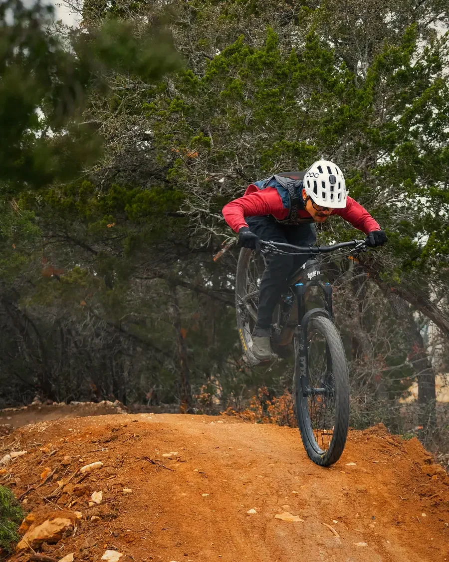 Rider airborne off a red-dirt jump lip with cedar trees behind, Texas Hill Country