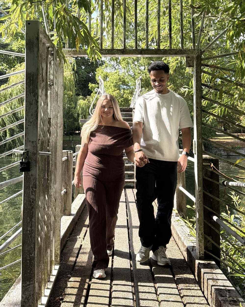 Jas and Josh holding hands as they walk across a sunlit footbridge.