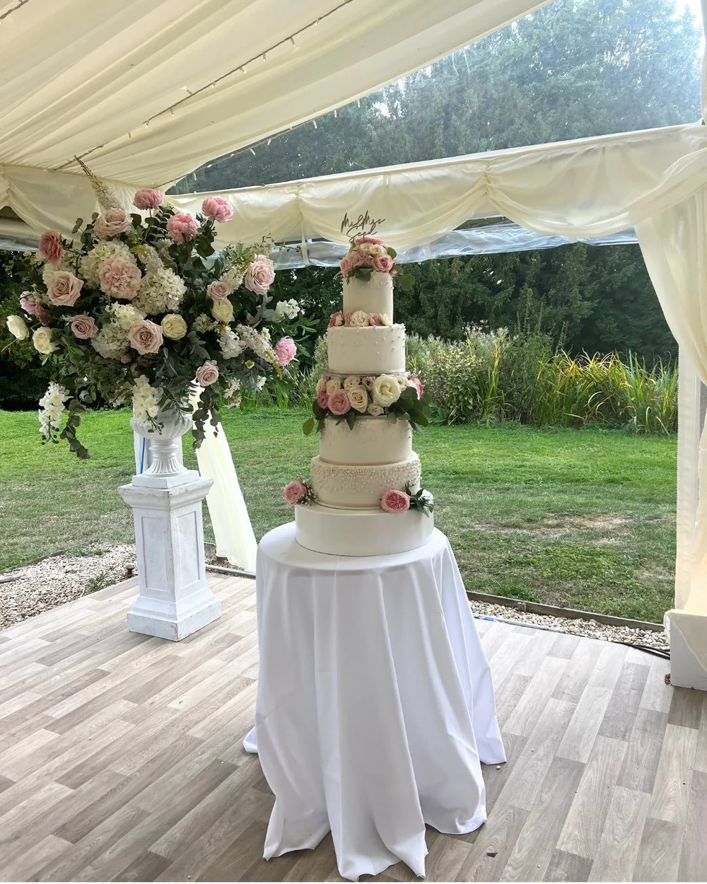 Ben and Colleen’s tiered white wedding cake with pink roses beside a floral arrangement.