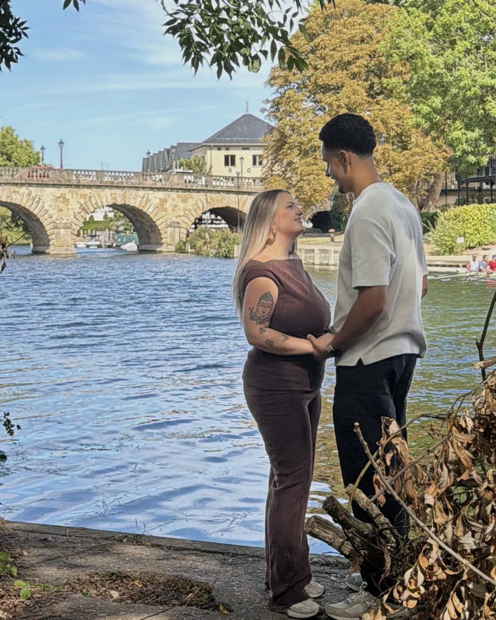 Jas and Josh holding hands by the river with a stone bridge behind them.