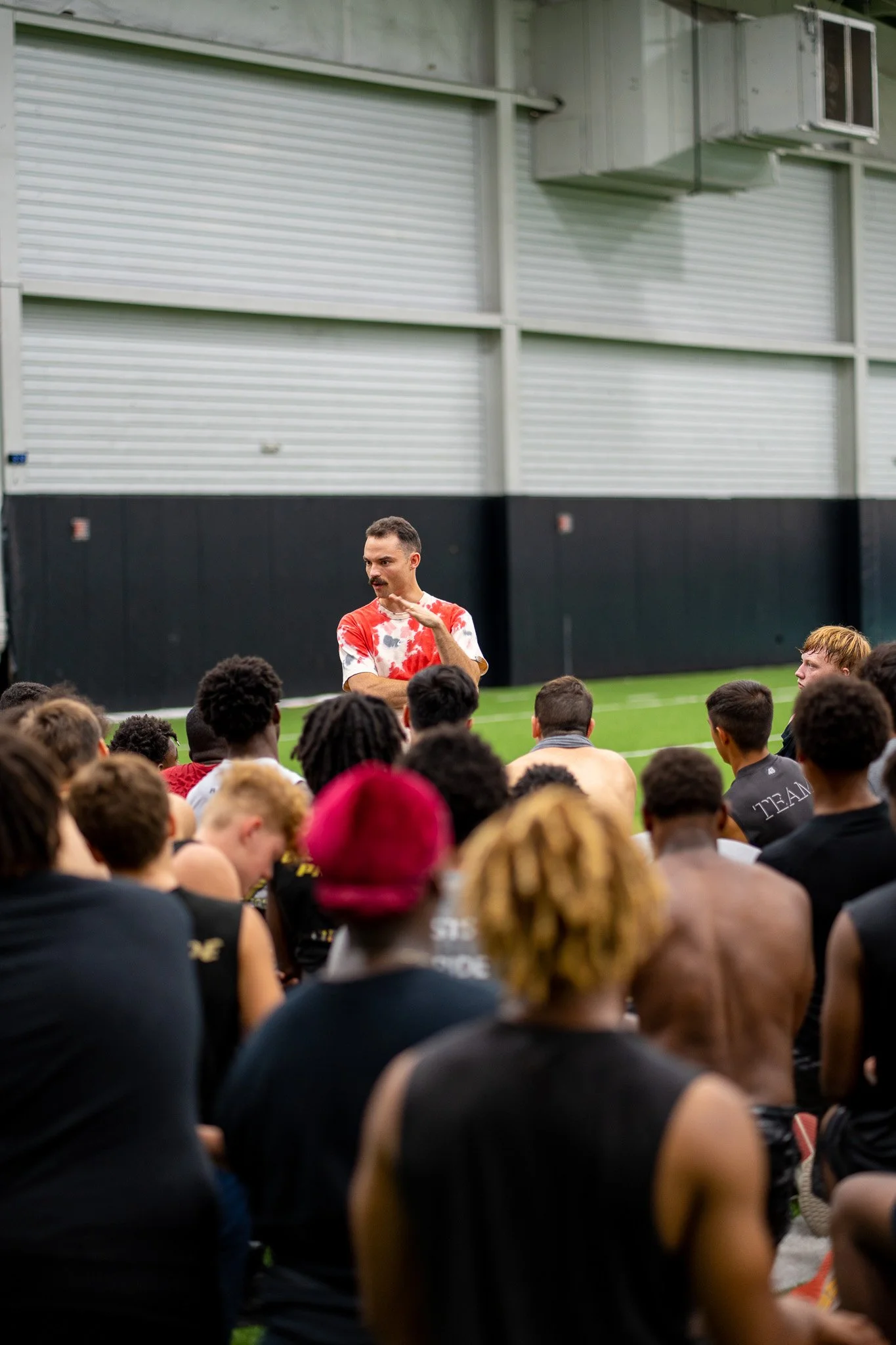 Seth in a red tie-dye shirt speaking with a football team gathered around him