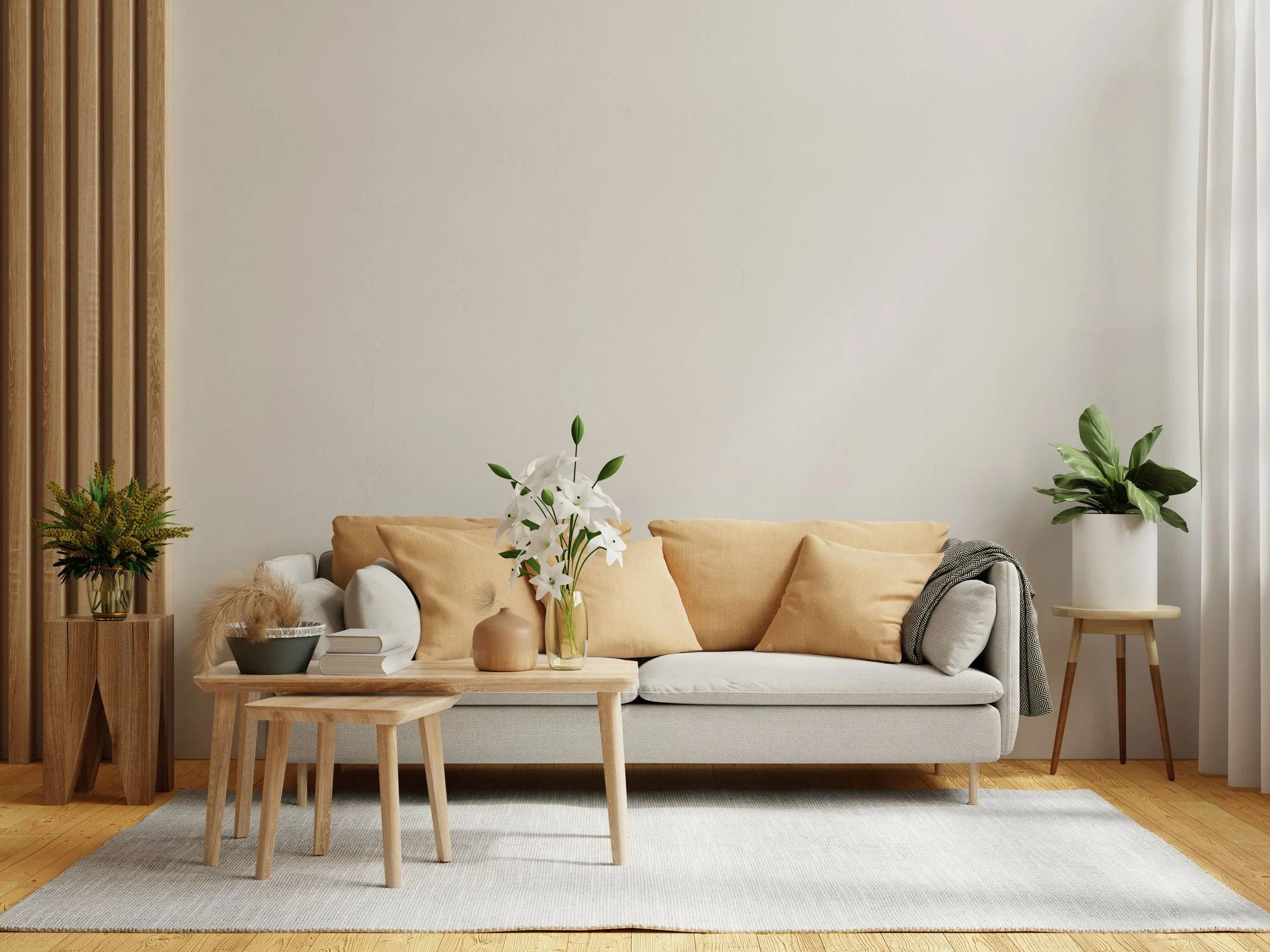 A pared-down neutral living room with a light sofa, soft gold pillows, simple wood coffee tables, one vase of white lilies and a few green plants