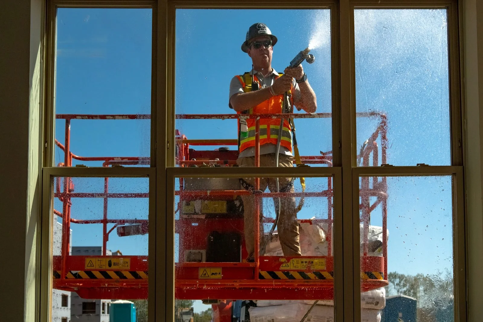 Green Home & Building technician on a lift spraying a calibrated water test across a window, seen from inside the building
