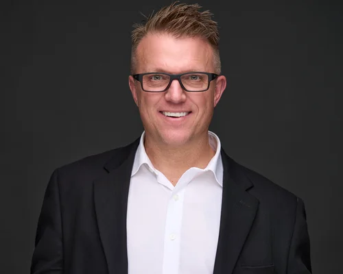 Headshot of a smiling man in glasses and a dark blazer against a dark backdrop, ATX Headshots