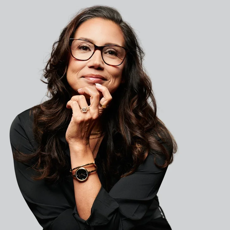 Woman with long dark wavy hair and glasses in a black blouse, hand at her chin, smiling on a light grey background. ATX Headshots, Austin.