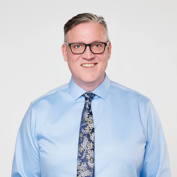 Headshot of a man in glasses, a light blue shirt and patterned tie with no jacket, light background