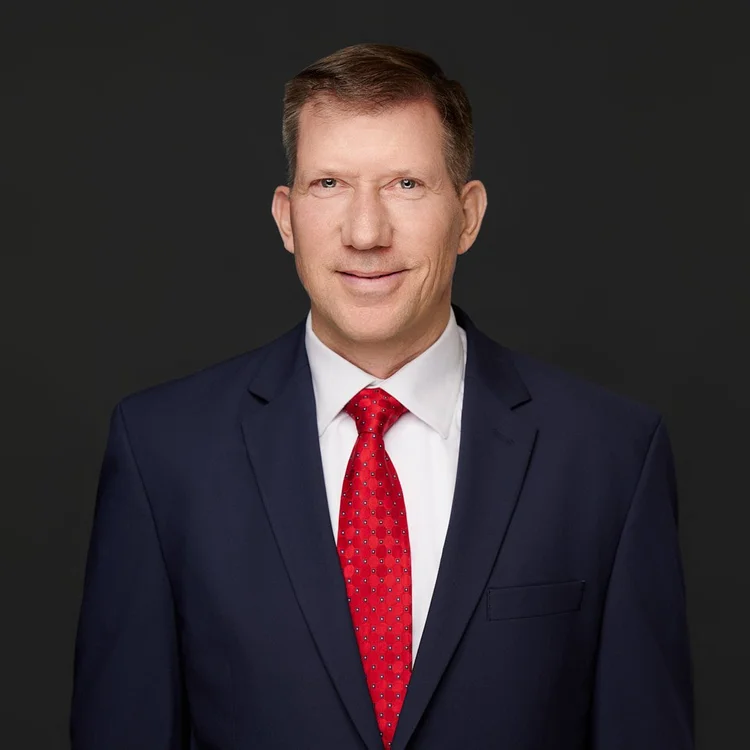 Headshot of a man in a navy suit, white shirt and red tie, dark background