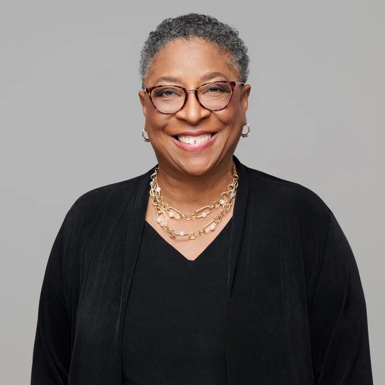 Woman with short grey hair and glasses in a black top and gold necklace, smiling on a grey background. ATX Headshots, Austin.