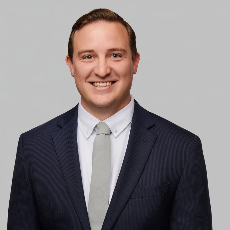 Attorney in a navy pinstripe suit and grey tie, smiling on a light grey background matched to his firm's team headshots. ATX Headshots, Austin.