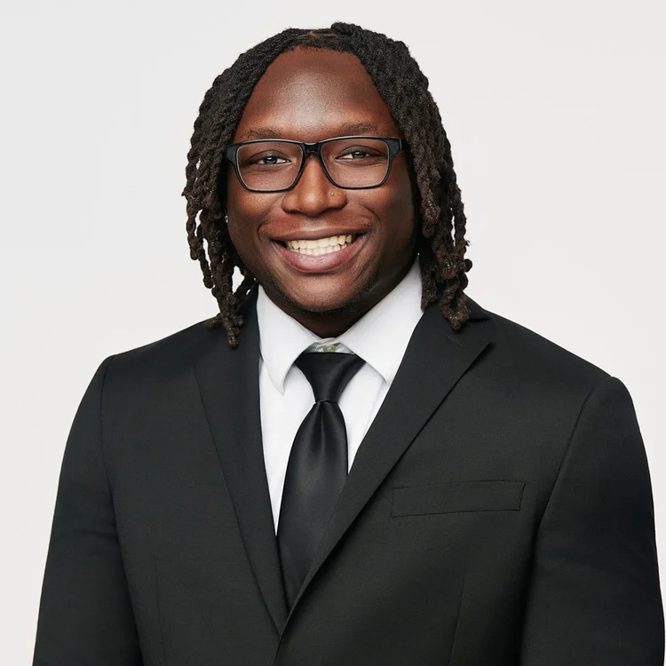 Headshot of a man in glasses, a black suit, white shirt and black tie, light background