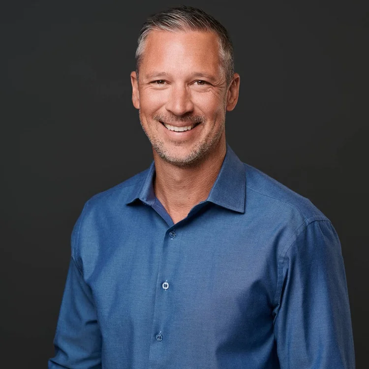 Man with greying hair in an open-collar blue shirt, smiling on a dark grey background. ATX Headshots, Austin.