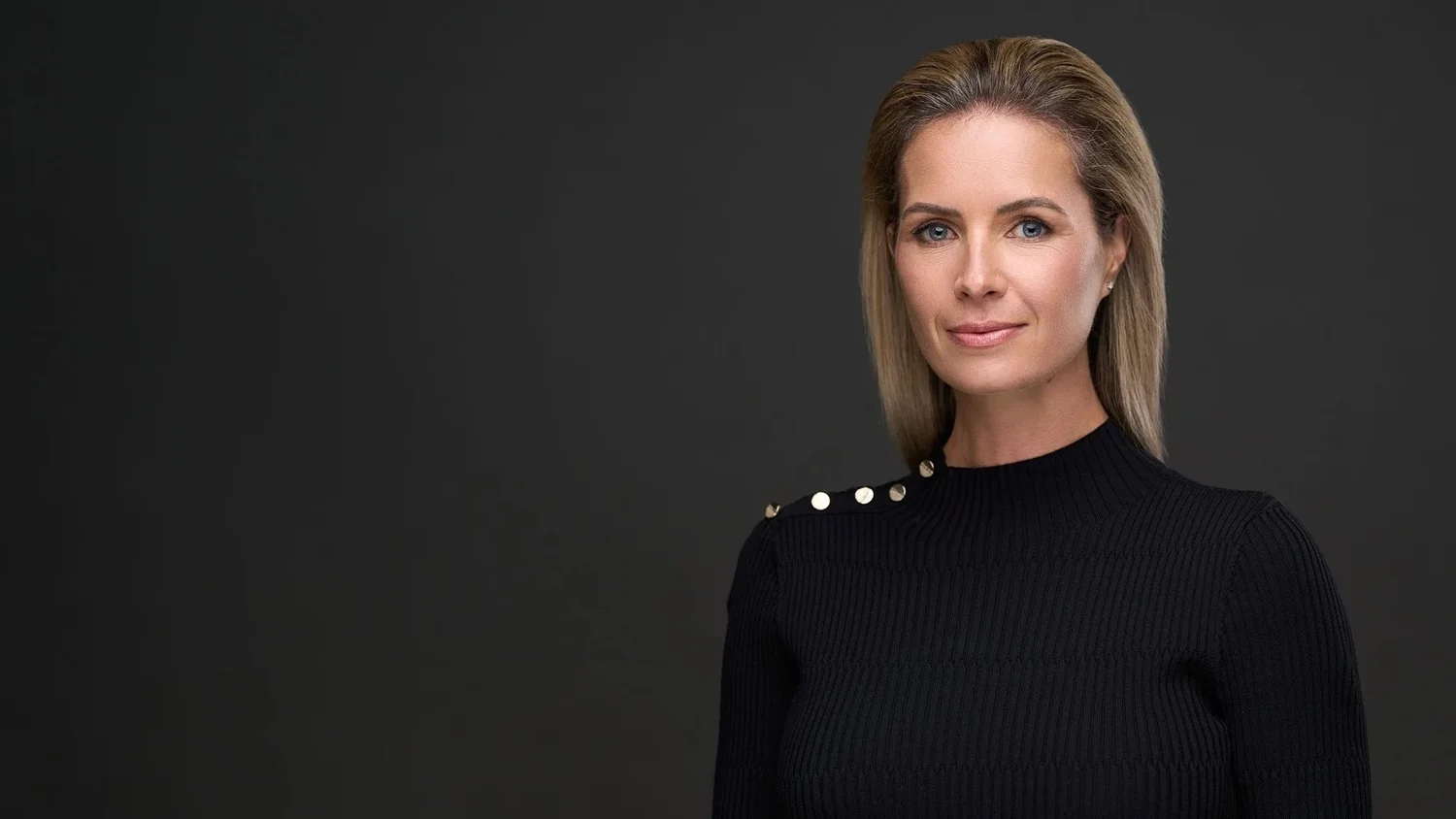 Headshot of a woman with shoulder-length blonde hair in a black ribbed top with gold buttons at the shoulder, on a dark grey background