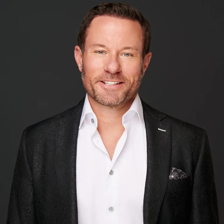 Man with a short beard in a black textured jacket and open white shirt, smiling on a dark background. ATX Headshots, Austin.