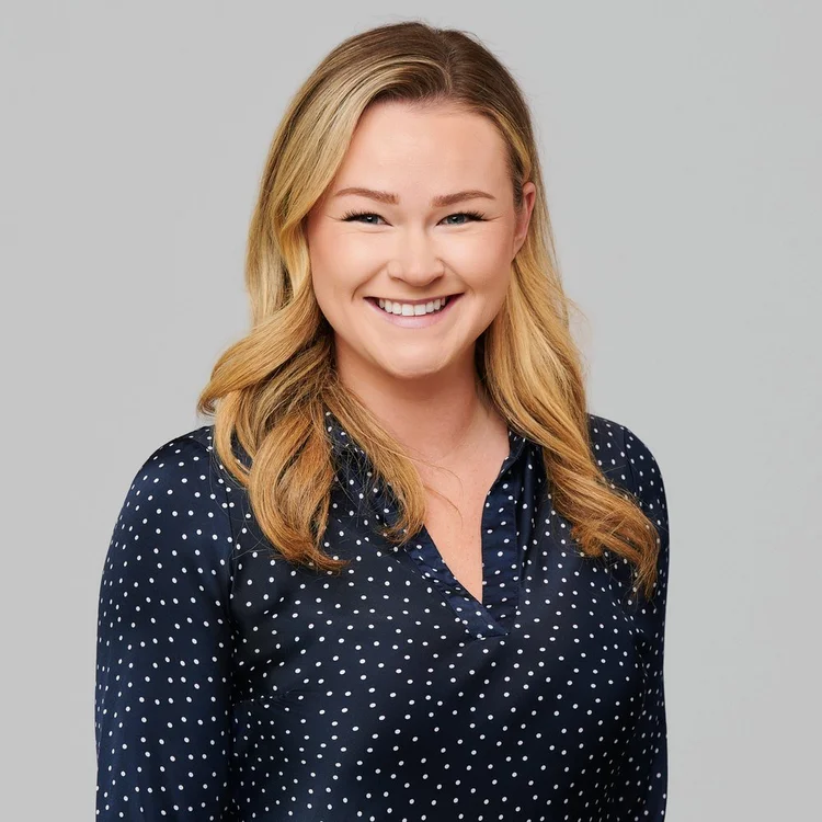 Woman with long wavy blonde hair in a navy polka-dot blouse, smiling on a light grey background. ATX Headshots, Austin.