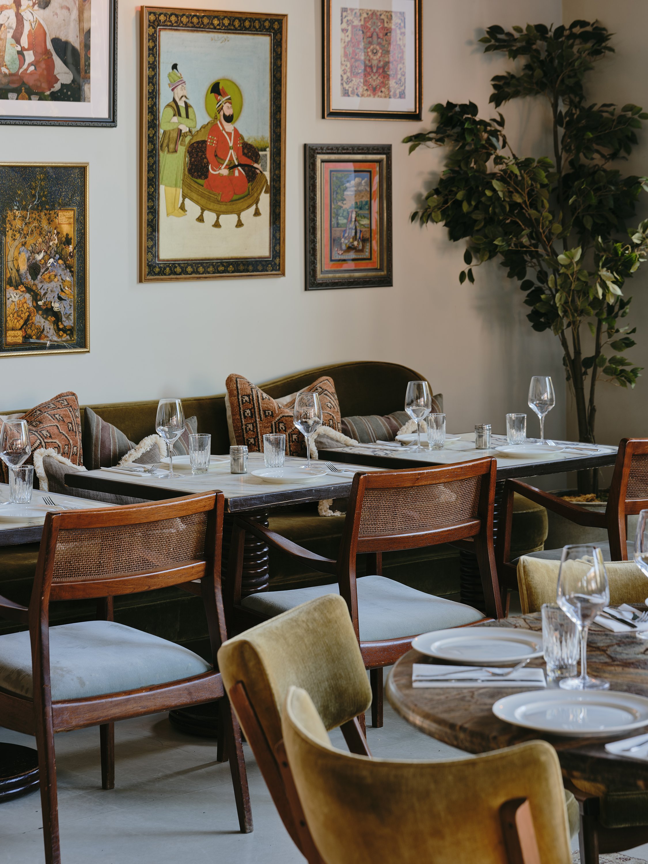 Interior of Berenjak at DUMBO House, featuring mid-century-inspired wooden furniture, cane chairs, and white table settings. The walls are adorned with colorful Persian artwork and soft lighting enhances the cozy atmosphere.