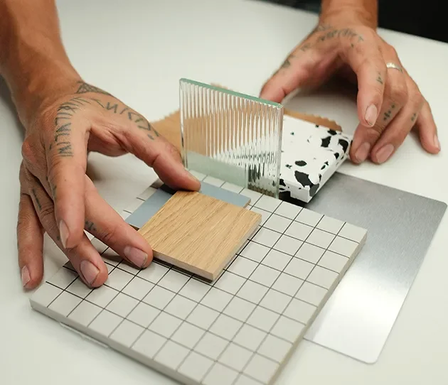 Hands arranging wood, terrazzo, metal and fluted glass material samples on a white tile with a grid pattern.